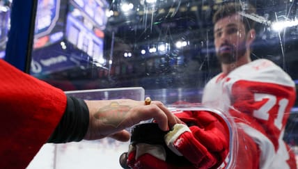 Dylan Larkin handing a puck through the boards while playing for the Detroit Red Wings but the hand grabbing it on the other side has a Minnesota Wild logo transposed on it.