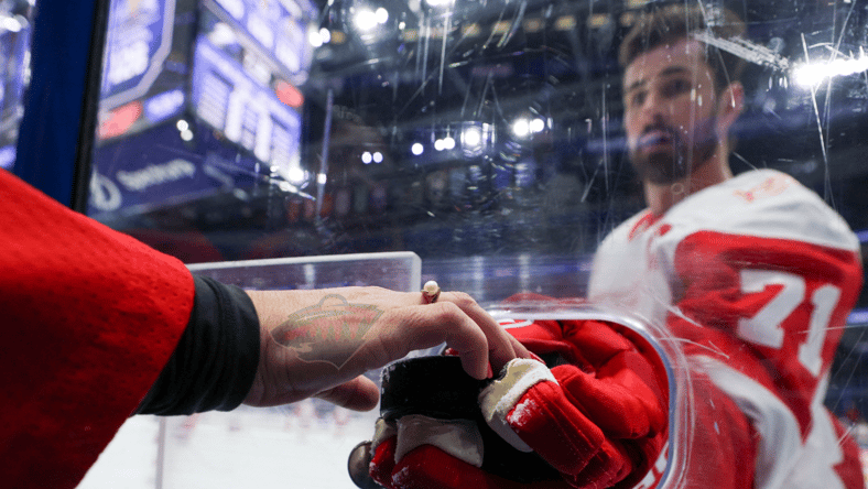 Dylan Larkin handing a puck through the boards while playing for the Detroit Red Wings but the hand grabbing it on the other side has a Minnesota Wild logo transposed on it.